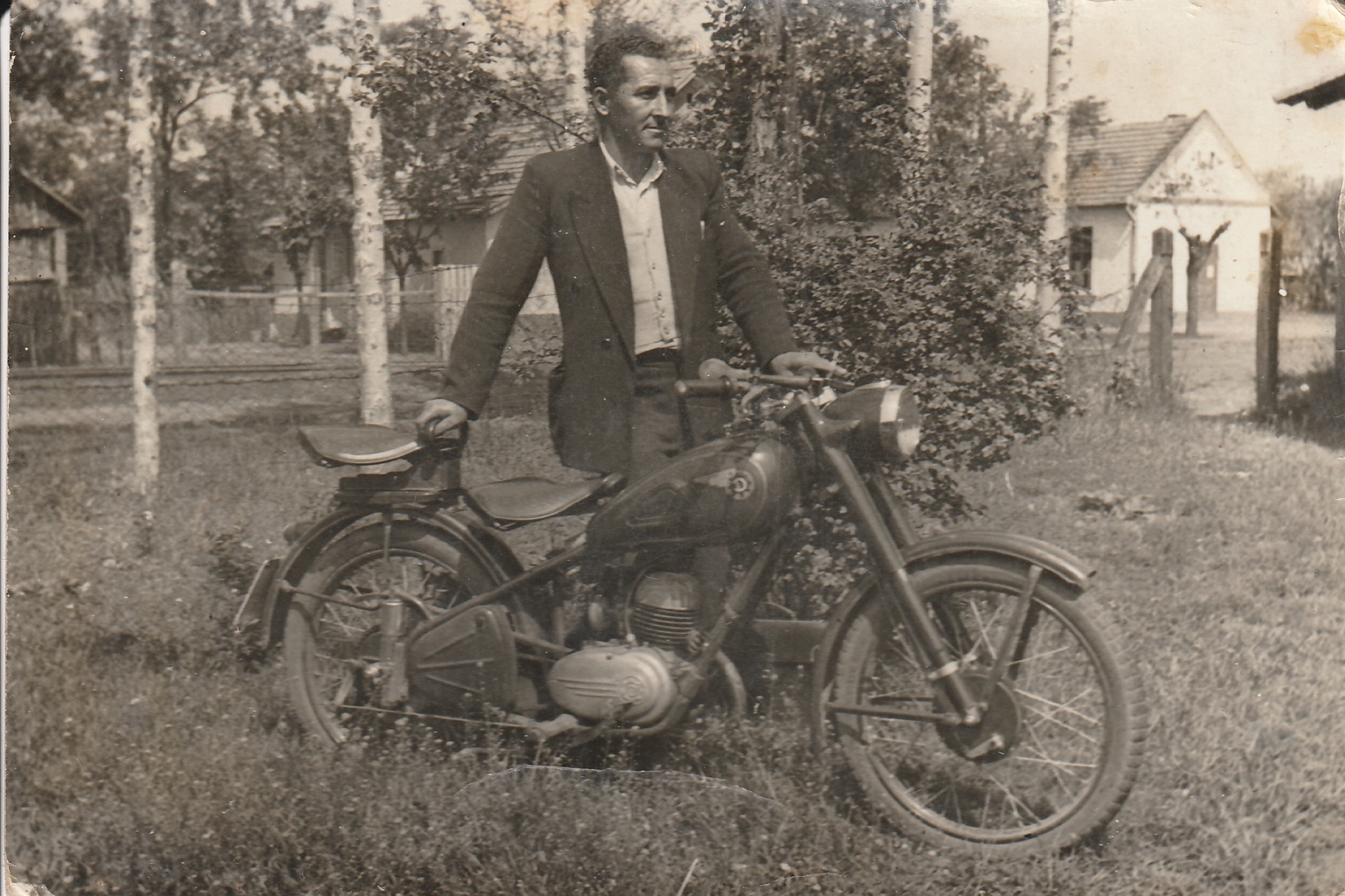 Régi fekete-fehér fotón férfi áll motorbicikli mellett, házak és fák között, poros, szemcsés háttérrel.