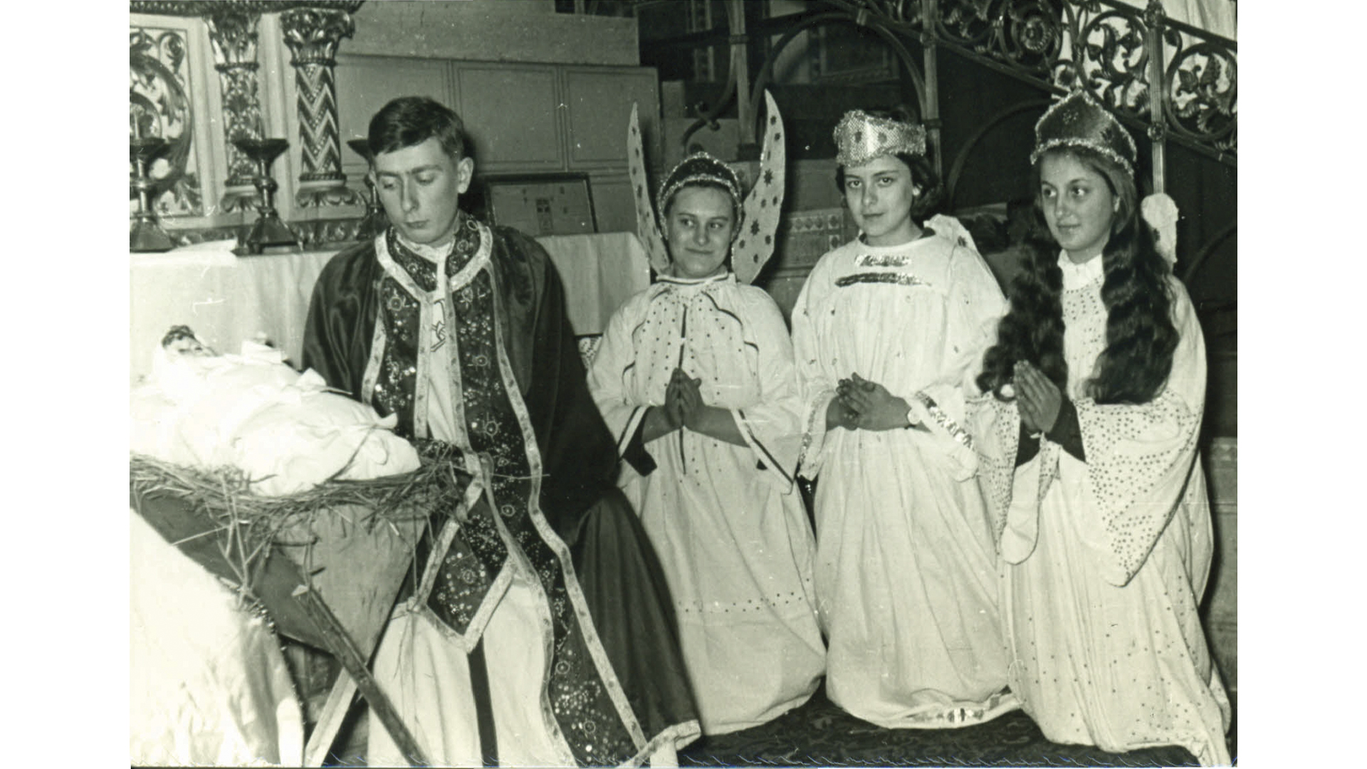 Négy fiatal ünnepi viseletben áll, fejdíszekkel és díszes ruhákkal, régi fotón, háttérben díszített ajtó.