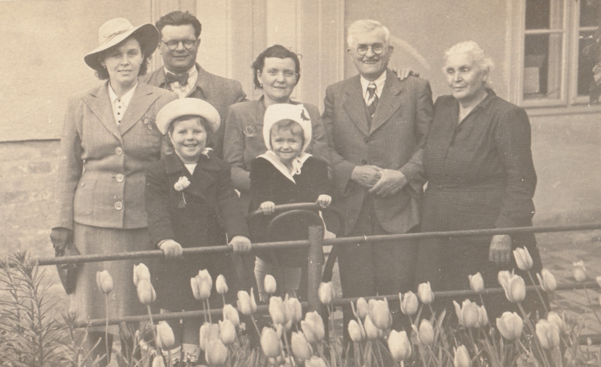 Régi fekete-fehér családi fotó: öt felnőtt és két kalapos gyermek virágokkal díszített kerítés előtt.