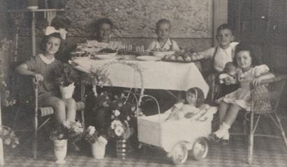 Régi családi fotó: ötenn az asztalnál ülnek, terített asztal, virágok és gyertyák, kerti háttérrel.